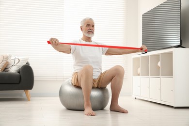 Senior man doing exercise with elastic resistance band on fitness ball at home