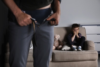 Photo of Man with unzipped pants standing near scared little boy on sofa indoors. Child in danger