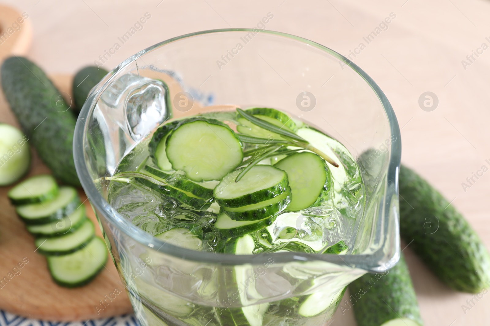 Photo of Refreshing cucumber water with rosemary in jug and vegetables on light table, closeup