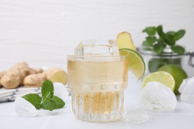 Glass of tasty ginger ale with ice cube and ingredients on white table, closeup