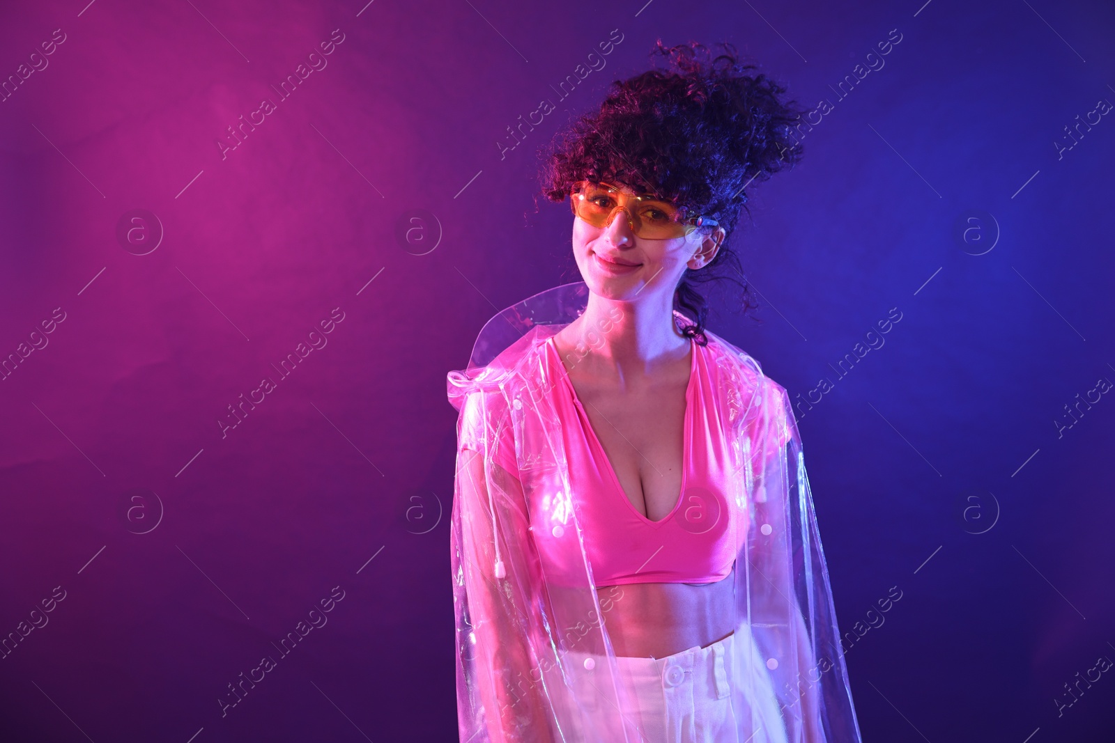 Photo of Beautiful young woman in transparent coat and sunglasses posing on color background in neon lights. Space for text