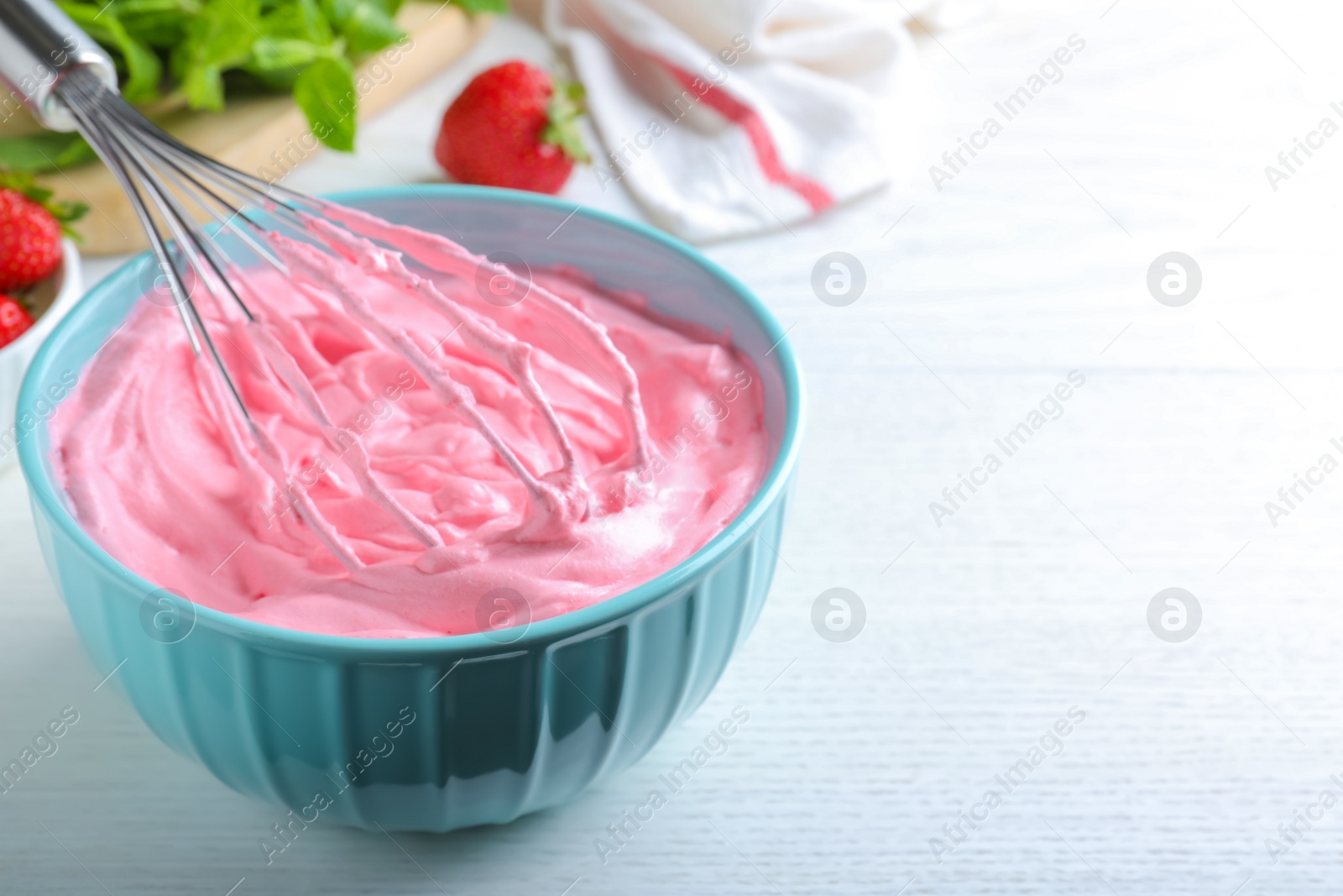 Photo of Whipping strawberry cream with balloon whisk on white wooden table, closeup. Space for text
