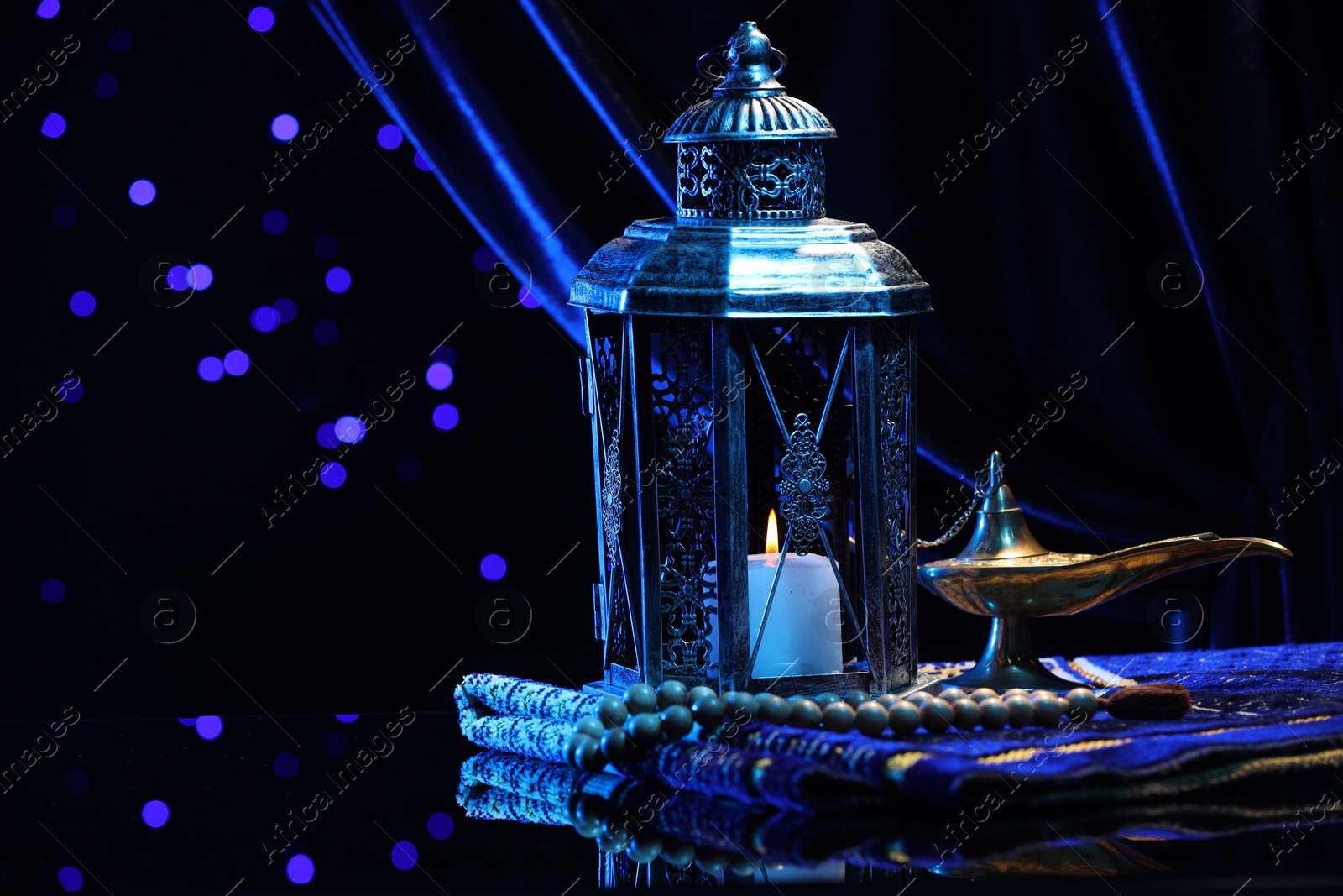 Photo of Arabic lantern, misbaha, Aladdin magic lamp and folded prayer mat on mirror surface against blurred lights at night. Space for text