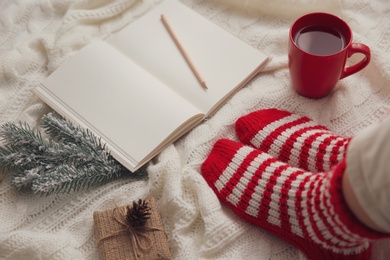 Photo of Woman with cup of hot winter drink and notebook on knitted plaid, closeup. Cozy season