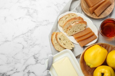 Delicious quince paste, bread, butter, cup of tea and fresh fruits on white marble table, flat lay. Space for text