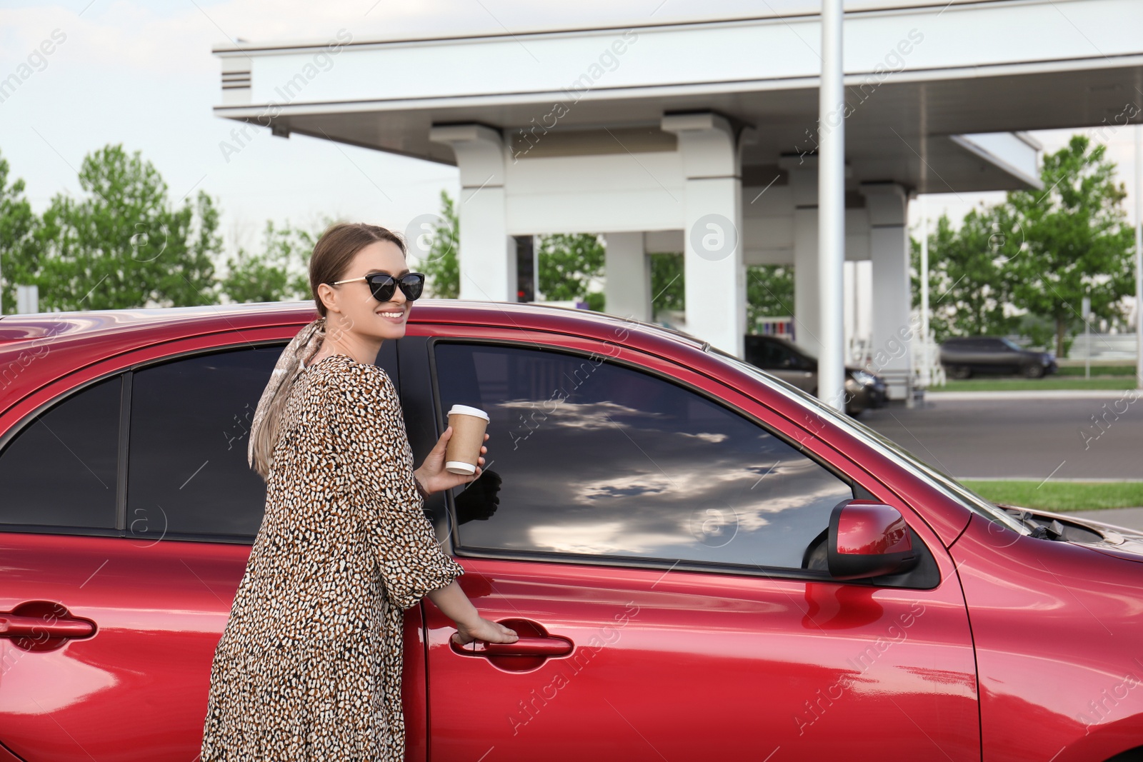 Photo of Beautiful young woman with coffee opening car door at gas station