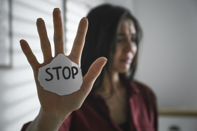 Photo of Crying young woman with sign STOP indoors, focus on hand. Domestic violence concept