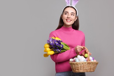 Photo of Happy woman in bunny ears headband holding wicker basket with painted Easter eggs and bouquet of flowers on grey background. Space for text