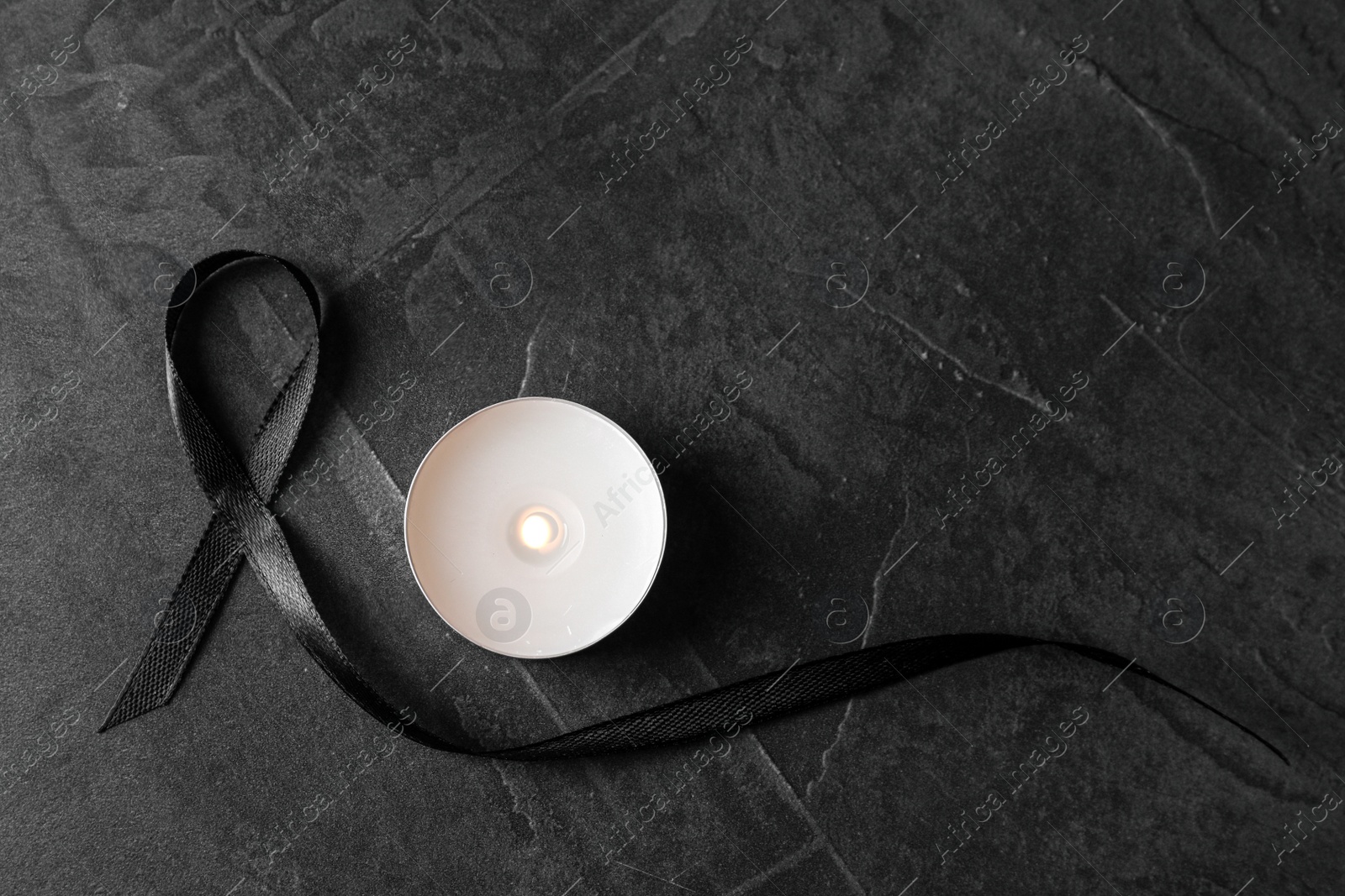 Photo of Black ribbon and burning candle on dark grey stone surface, top view. Funeral symbols