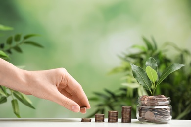 Woman putting coin onto stack, glass jar and green plant on white table against blurred background, closeup. Space for text