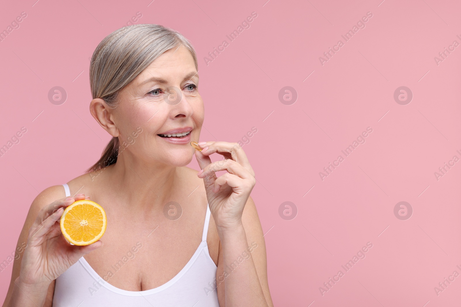 Photo of Beautiful woman with half of orange taking vitamin capsule on pink background, space for text