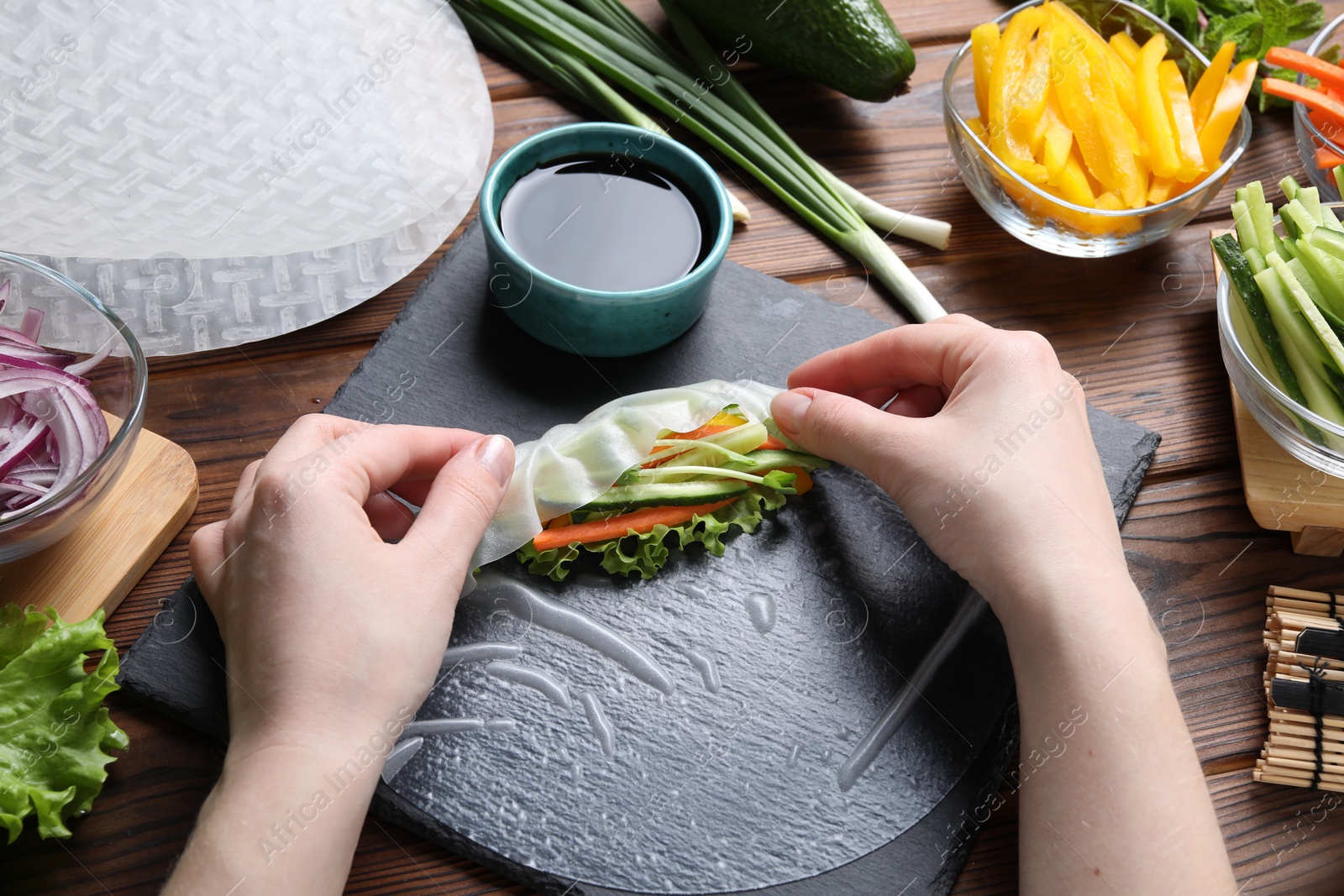 Photo of Woman wrapping spring roll at wooden table with products, closeup