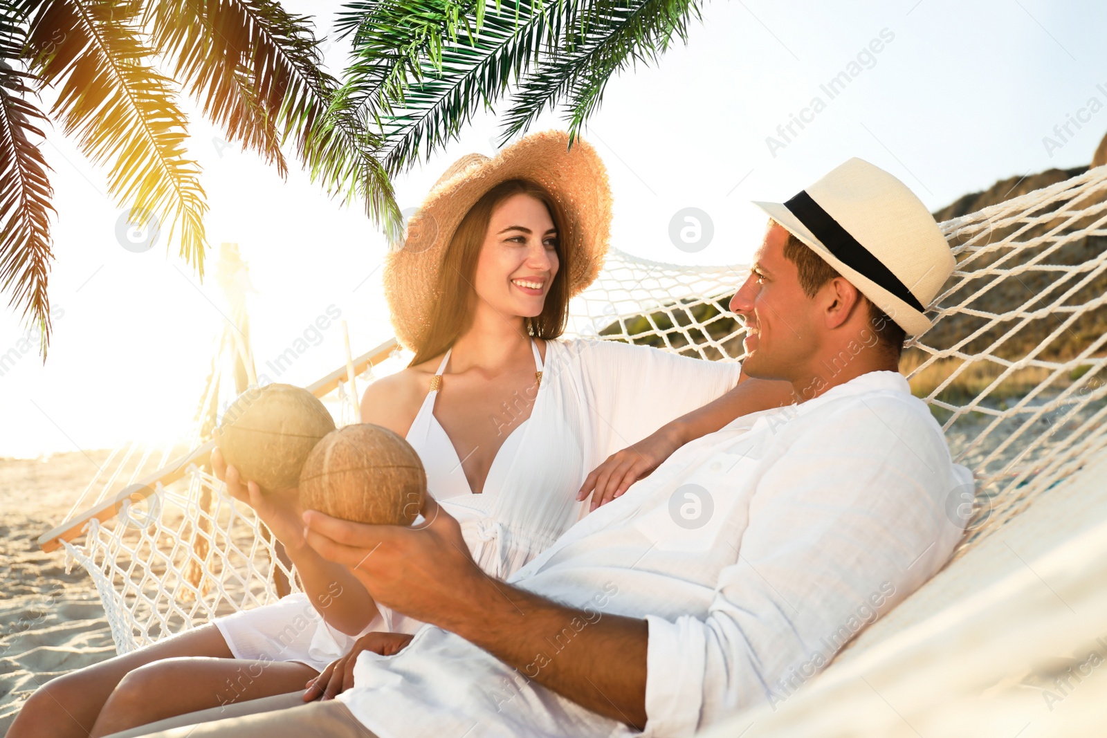 Image of Couple with exotic cocktails relaxing in hammock under green palm leaves on beach