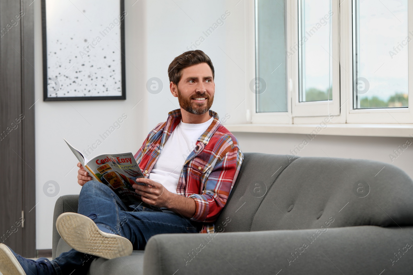 Photo of Smiling bearded man with magazine on sofa at home