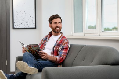 Smiling bearded man with magazine on sofa at home