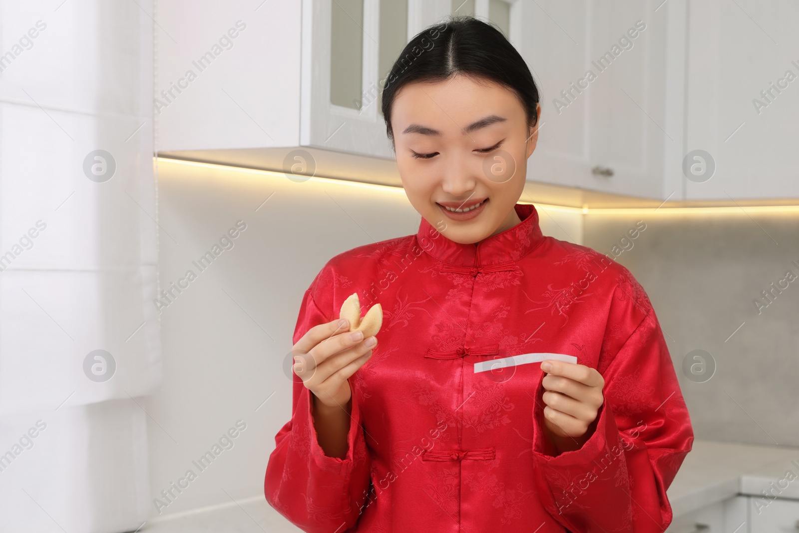 Photo of Asian woman holding tasty fortune cookie with prediction in kitchen. Space for text