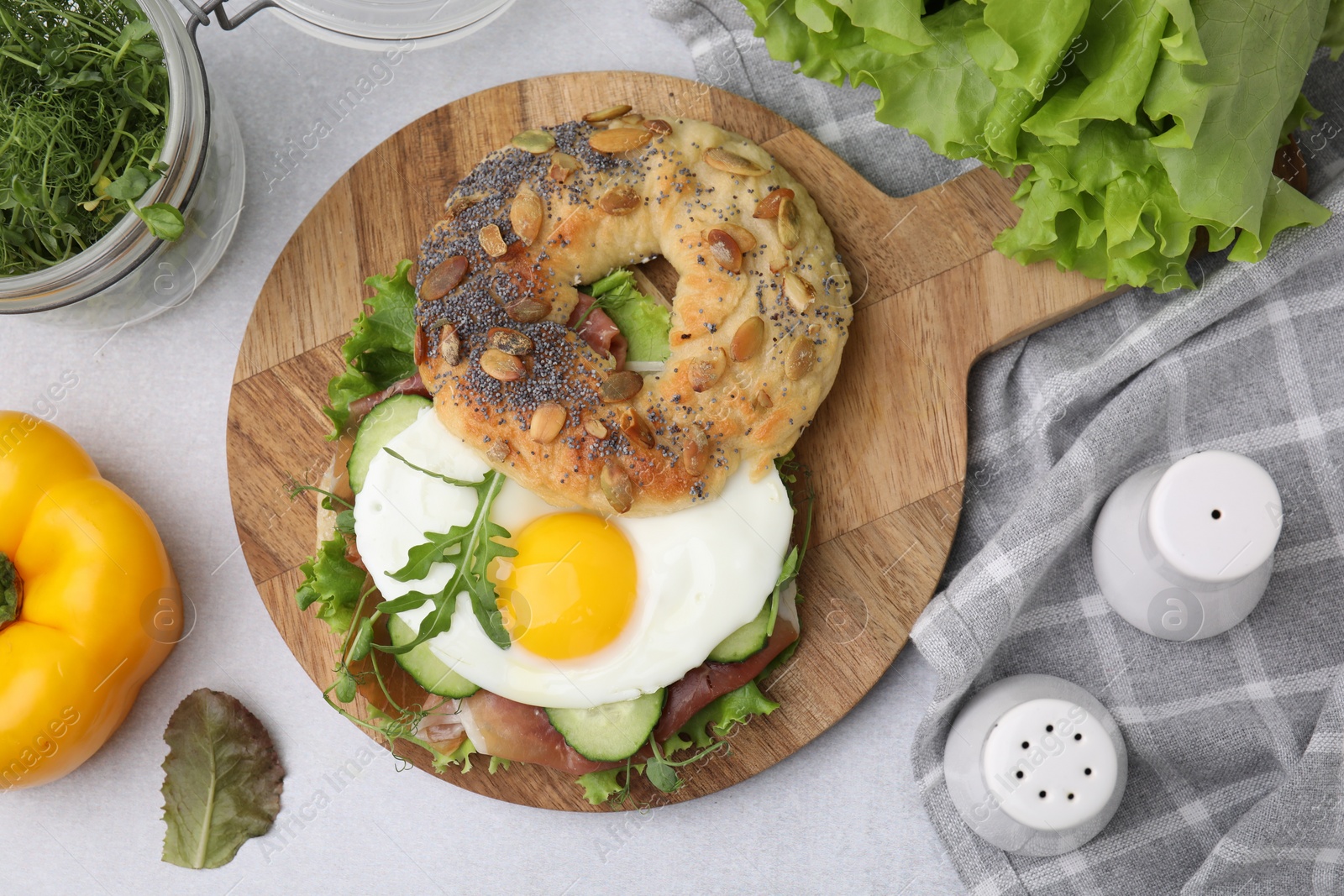 Photo of Tasty bagel with cured ham, egg, cucumber and salad mix on light table, flat lay