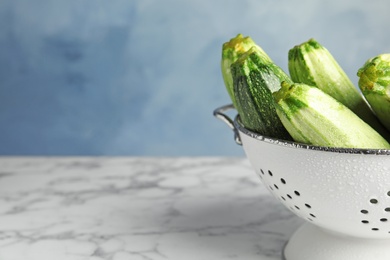Photo of Colander with fresh ripe green zucchinis on marble table against blue background, space for text