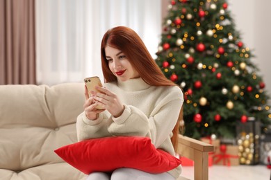 Photo of Beautiful young woman using smartphone on sofa near Christmas tree at home