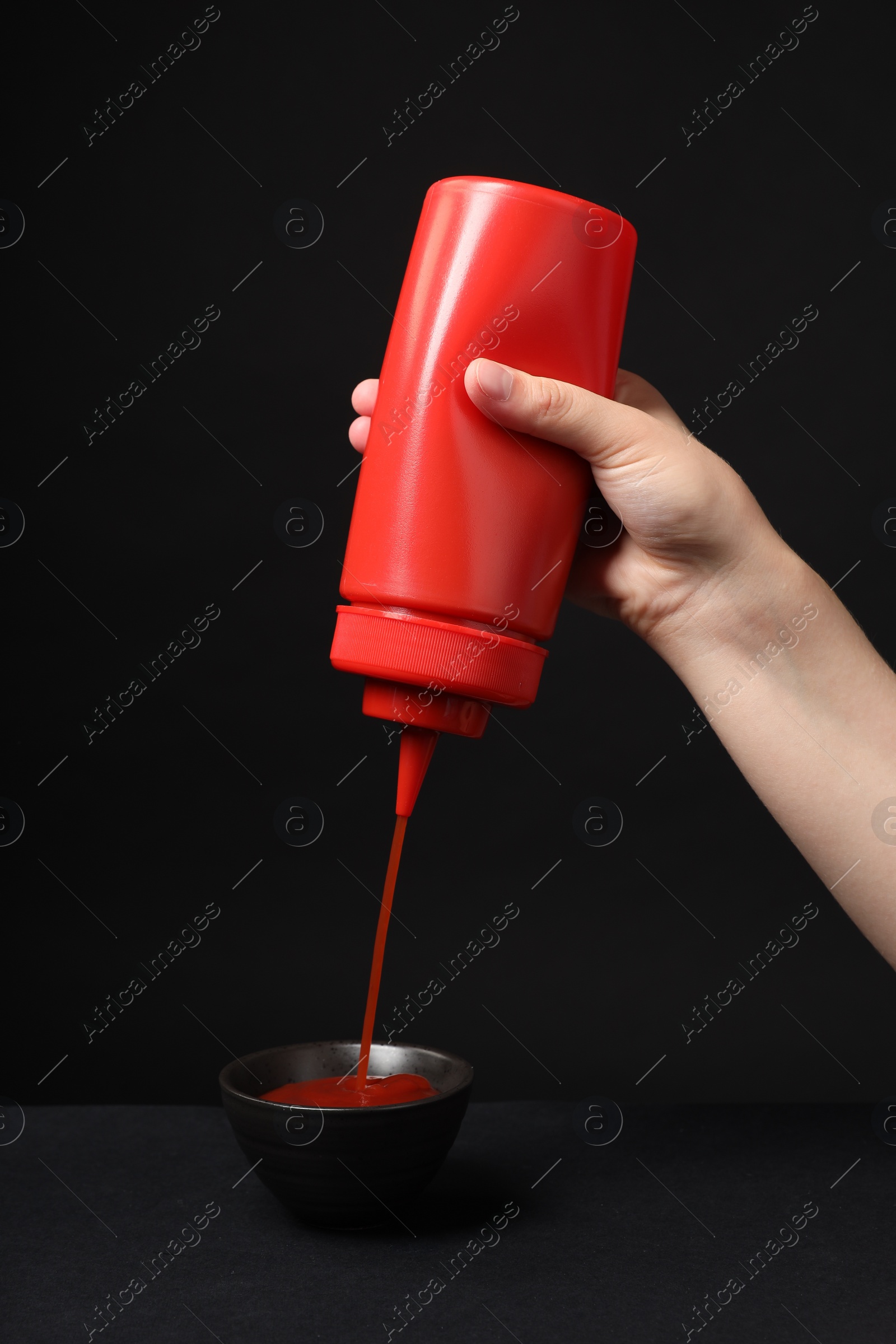 Photo of Woman pouring tasty ketchup from bottle into bowl against black background, closeup