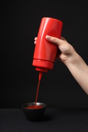 Woman pouring tasty ketchup from bottle into bowl against black background, closeup