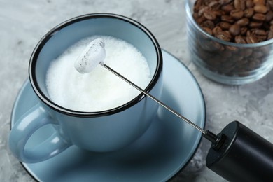 Photo of Mini mixer (milk frother), whipped milk in cup and coffee beans on grey textured table, closeup