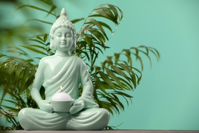 Buddhism religion. Decorative Buddha statue with burning candle on table and houseplant against turquoise wall, space for text