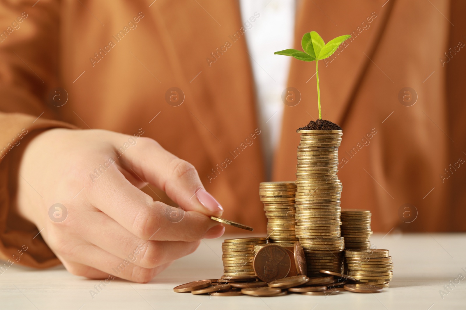 Photo of Woman putting coin onto stack with green sprout at white table, closeup. Investment concept