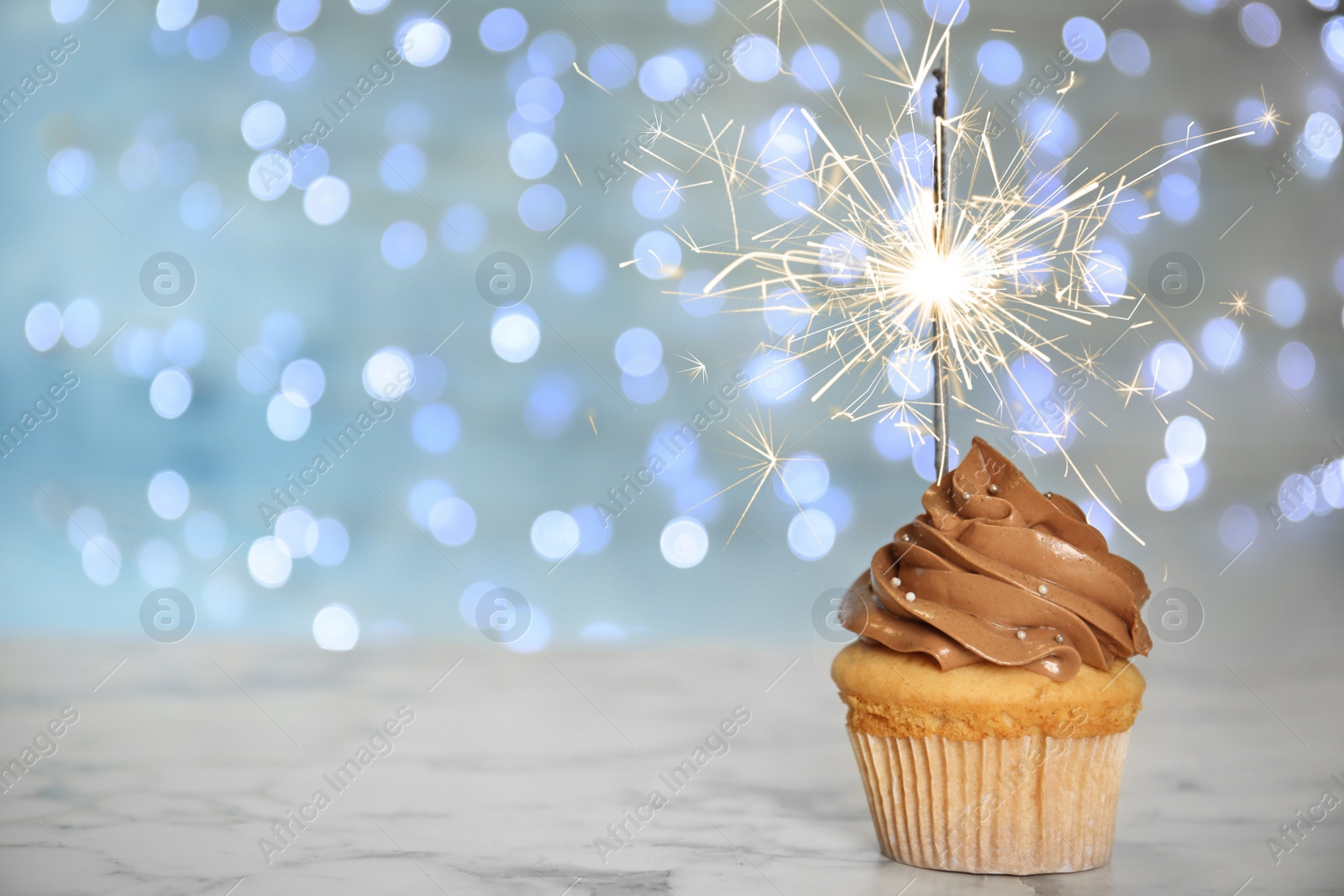 Image of Birthday cupcake with sparkler on table against blurred lights. Space for text