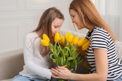 Happy mother and her cute daughter with bouquet of yellow tulips on sofa at home