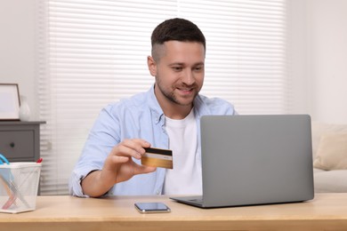 Handsome man with credit card using laptop for online shopping at wooden table in room