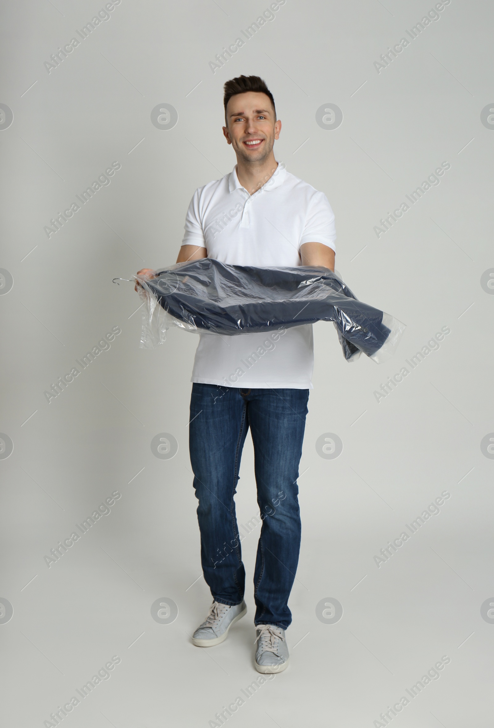 Photo of Man holding hanger with jacket in plastic bag on light grey background. Dry-cleaning service