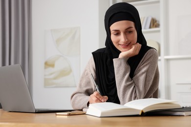 Muslim woman writing notes near laptop at wooden table in room