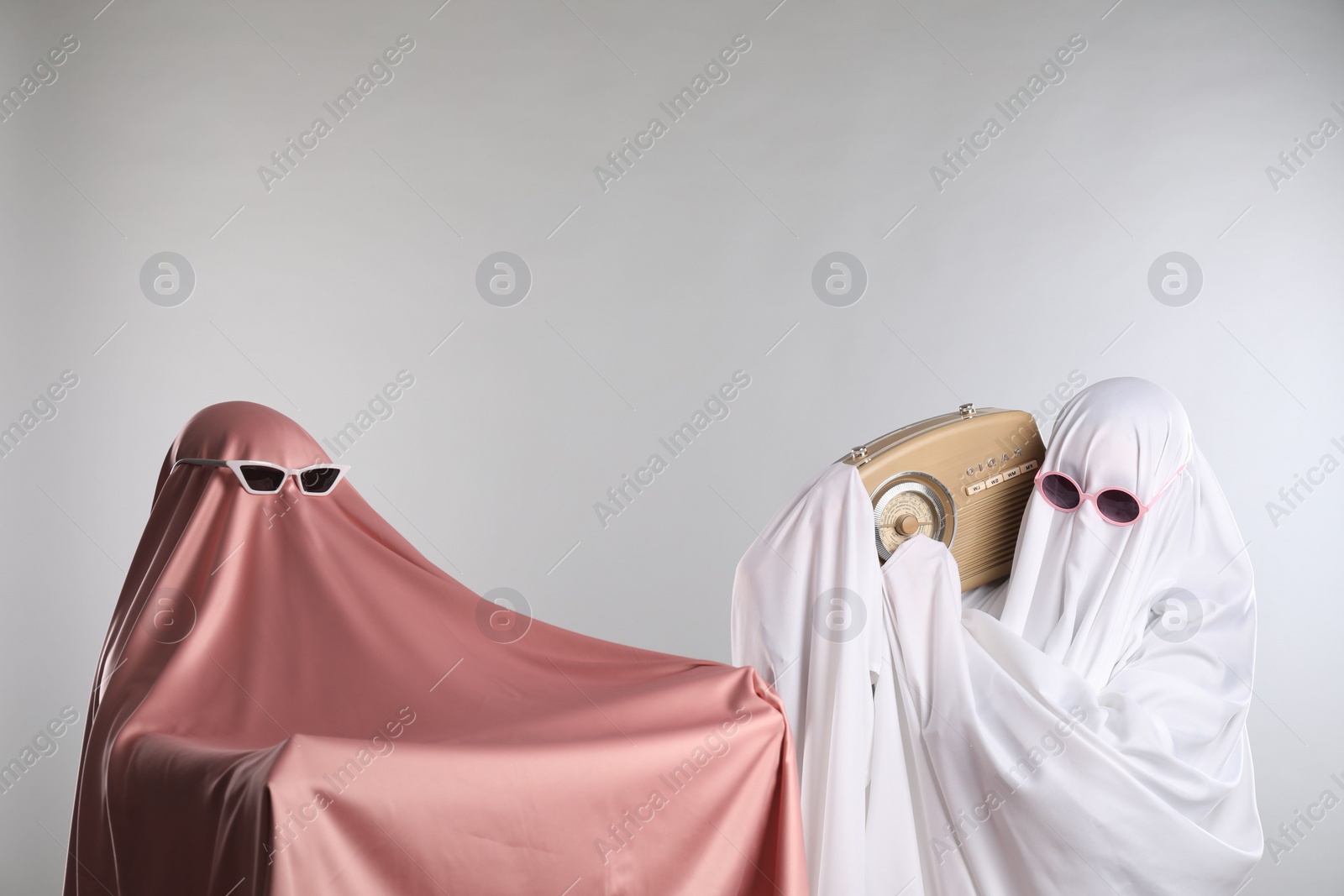 Photo of Glamorous ghosts. Women in color sheets and sunglasses with retro radio receiver on light grey background