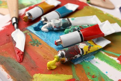 Photo of Tubes of colorful oil paints and spatula on canvas with abstract painting, closeup