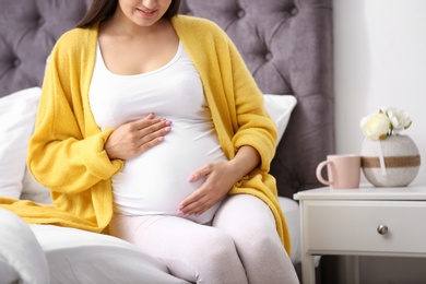 Young pregnant woman sitting on bed and touching her belly at home, closeup