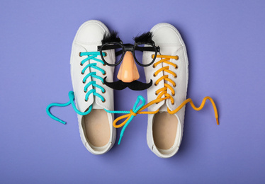 Shoes tied together and funny glasses on lilac background, flat lay. April Fool's Day