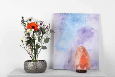 Photo of Stylish ikebana as house decor. Beautiful fresh flowers and eucalyptus branch on table near picture and salt lamp indoors