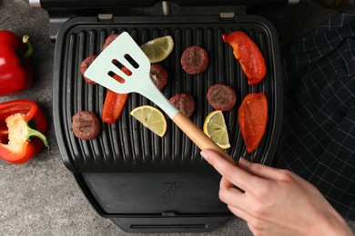 Photo of Woman cooking meat balls with bell peppers and lemon on electric grill at grey textured table, top view