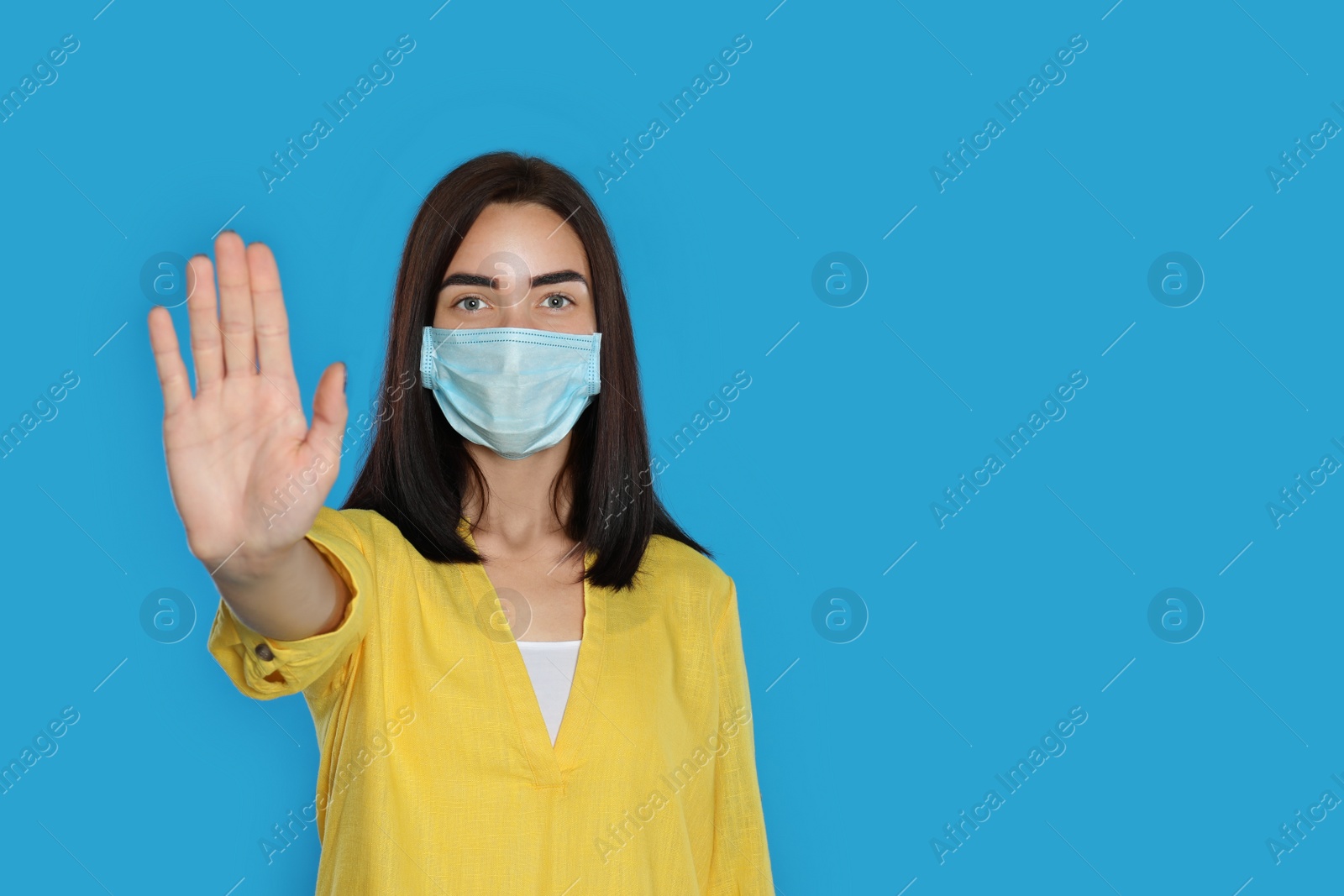 Photo of Young woman in protective mask showing stop gesture on light blue background, space for text. Prevent spreading of coronavirus