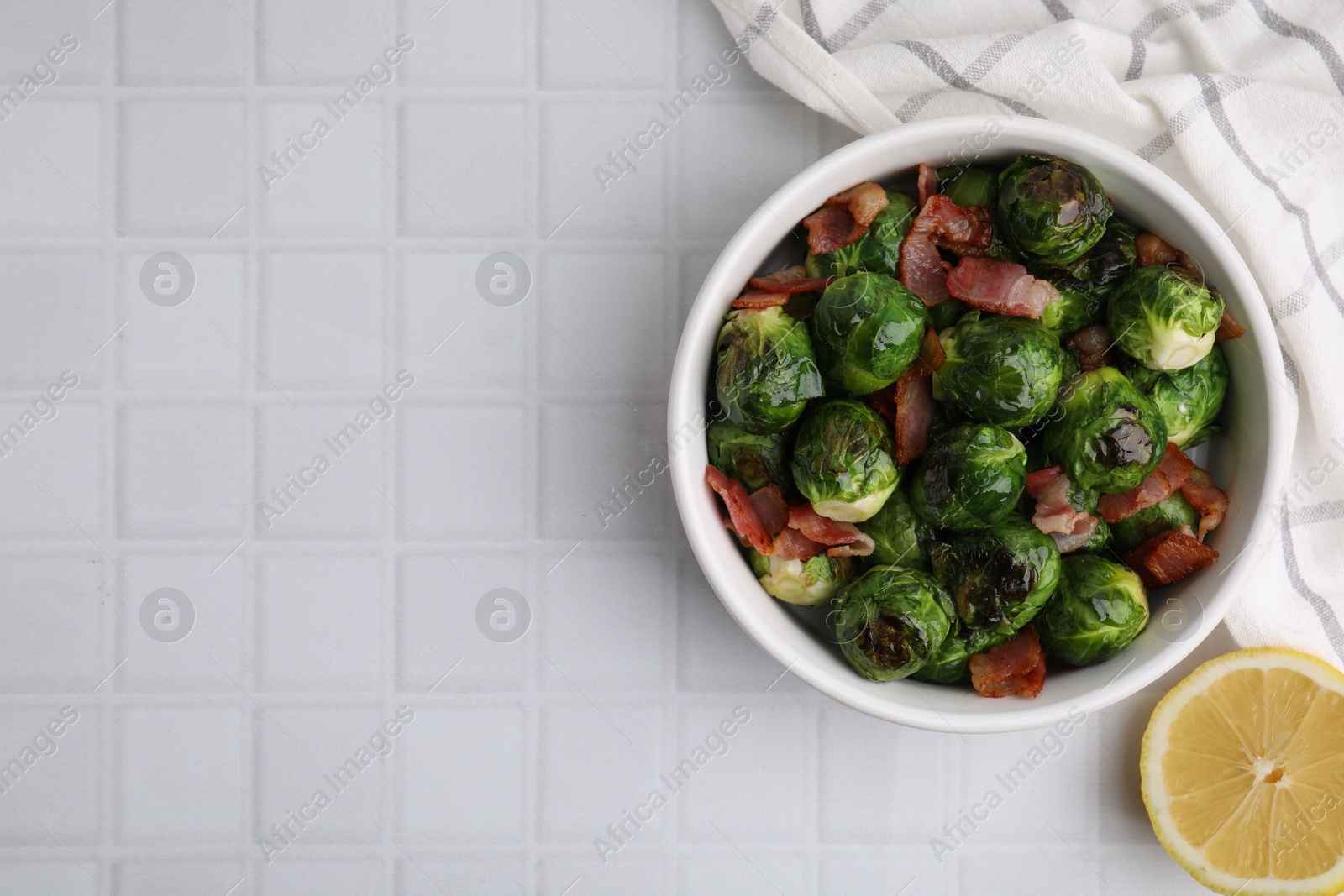 Photo of Delicious roasted Brussels sprouts, bacon and lemon on white tiled table, top view. Space for text