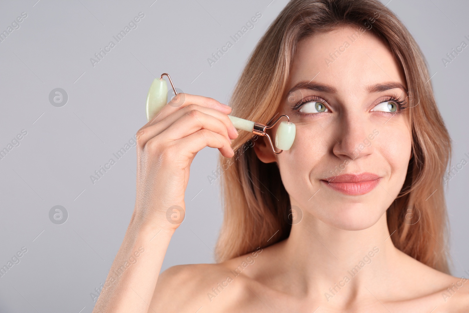 Photo of Young woman using natural jade face roller on light grey background