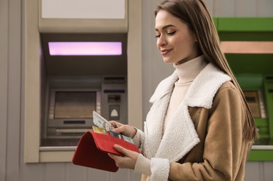 Woman putting money into wallet near cash machine outdoors