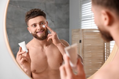 Photo of Handsome man applying moisturizing cream onto his face near mirror in bathroom
