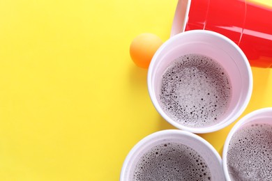 Plastic cups and ball on yellow background, flat lay with space for text. Beer pong game