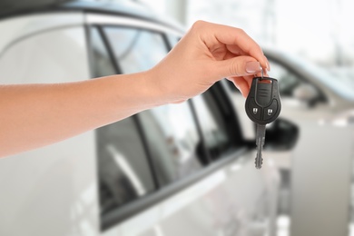 Car buying. Woman holding key against blurred automobile, closeup
