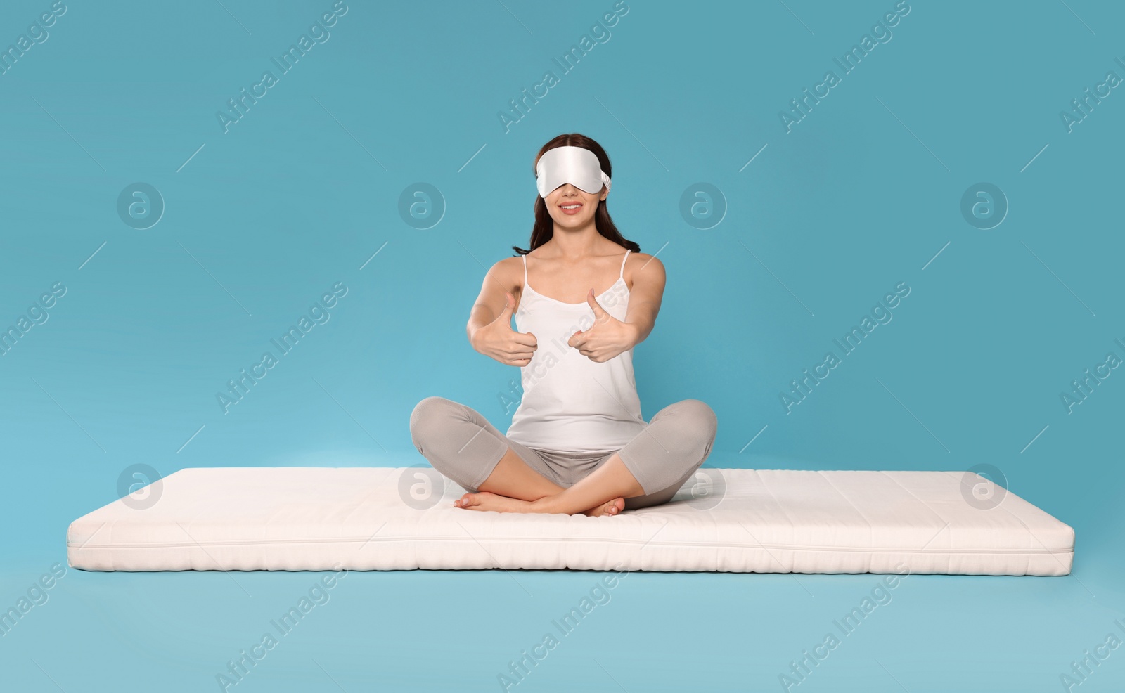 Photo of Woman in sleep mask sitting on soft mattress and showing thumbs up against light blue background