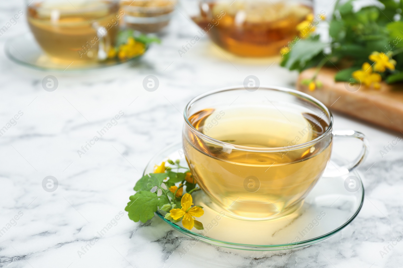 Photo of Glass cup of aromatic celandine tea and flowers on white marble table, space for text