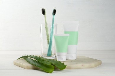 Tubes of toothpaste, toothbrushes and fresh aloe vera leaves on white wooden table