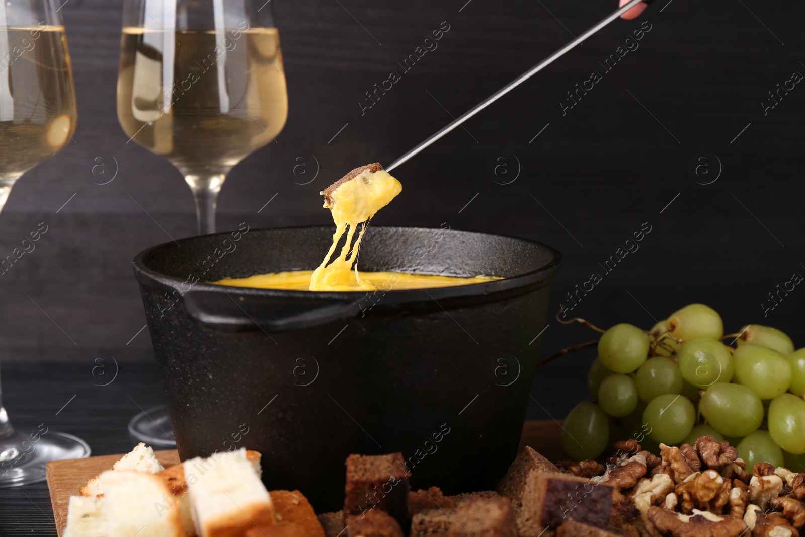 Photo of Dipping piece of bread into fondue pot with melted cheese on table, closeup